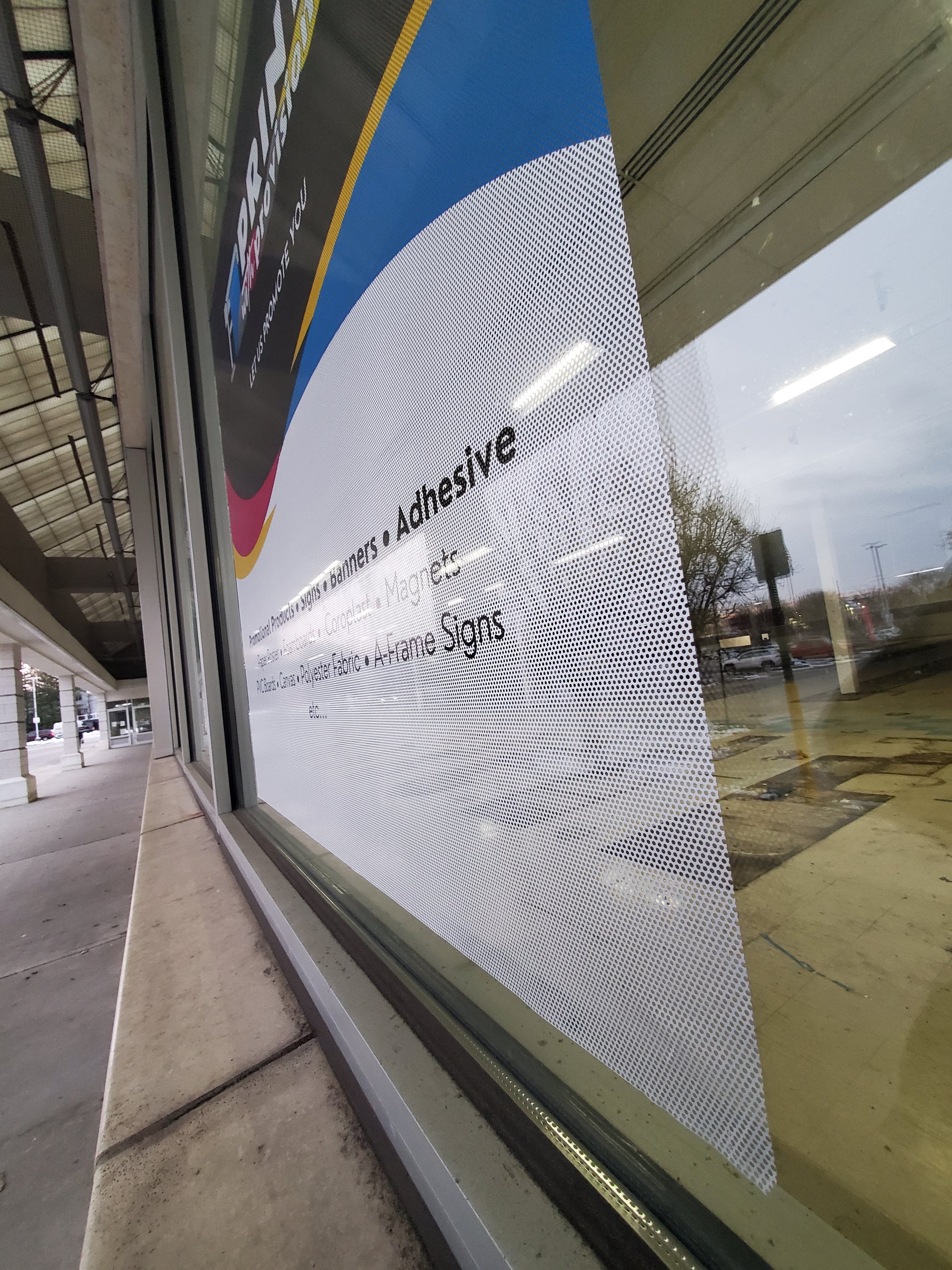 Custom perforated window film with Print With Provision branding, viewed from outside at an angle, installed on storefront window for promotional advertising.